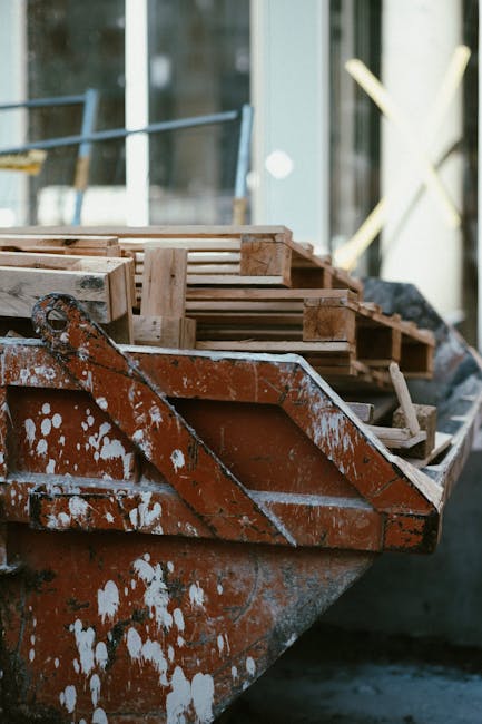 A large, open-top wooden skip with visibly weathered, dark brown planks is positioned on a wet pavement or road in an outdoor urban setting during nighttime. The skip is partially filled with various 