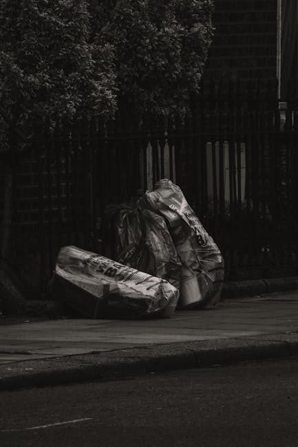 A large collection of waste materials and rubbish bags is piled around a communal litter bin on a paved urban street. The waste includes black and black-and-white plastic rubbish bags, brown cardboard