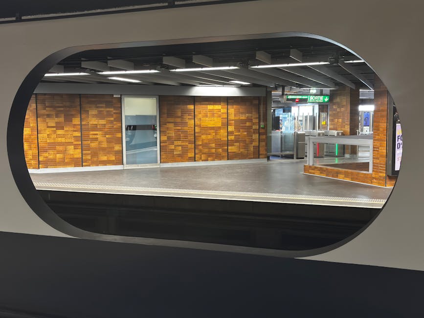 View through an oval-shaped window of a modern underground station platform featuring wooden panelled walls with a warm, natural wood finish. The platform has a smooth, light grey floor with a tactile